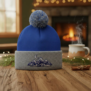Blue and gray knit beanie with mountain design, pom pom, on rustic table by cozy fireplace