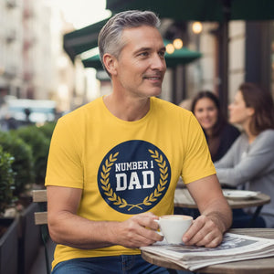 Smiling man in yellow Number 1 Dad t-shirt enjoying coffee outdoors at a café table.