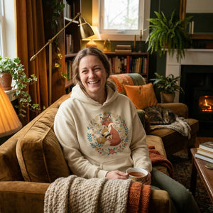 Smiling woman in cozy living room wearing cream hoodie with fox graphic, holding a cup of tea