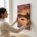 Woman hanging canvas wall art of a sunset lake scene with a boat and trees.