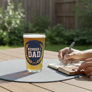 Pint glass with 'Number 1 Dad' design on outdoor table beside hands writing in a notebook