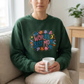 Woman wearing green World's Best Mom sweatshirt holding a mug on a couch